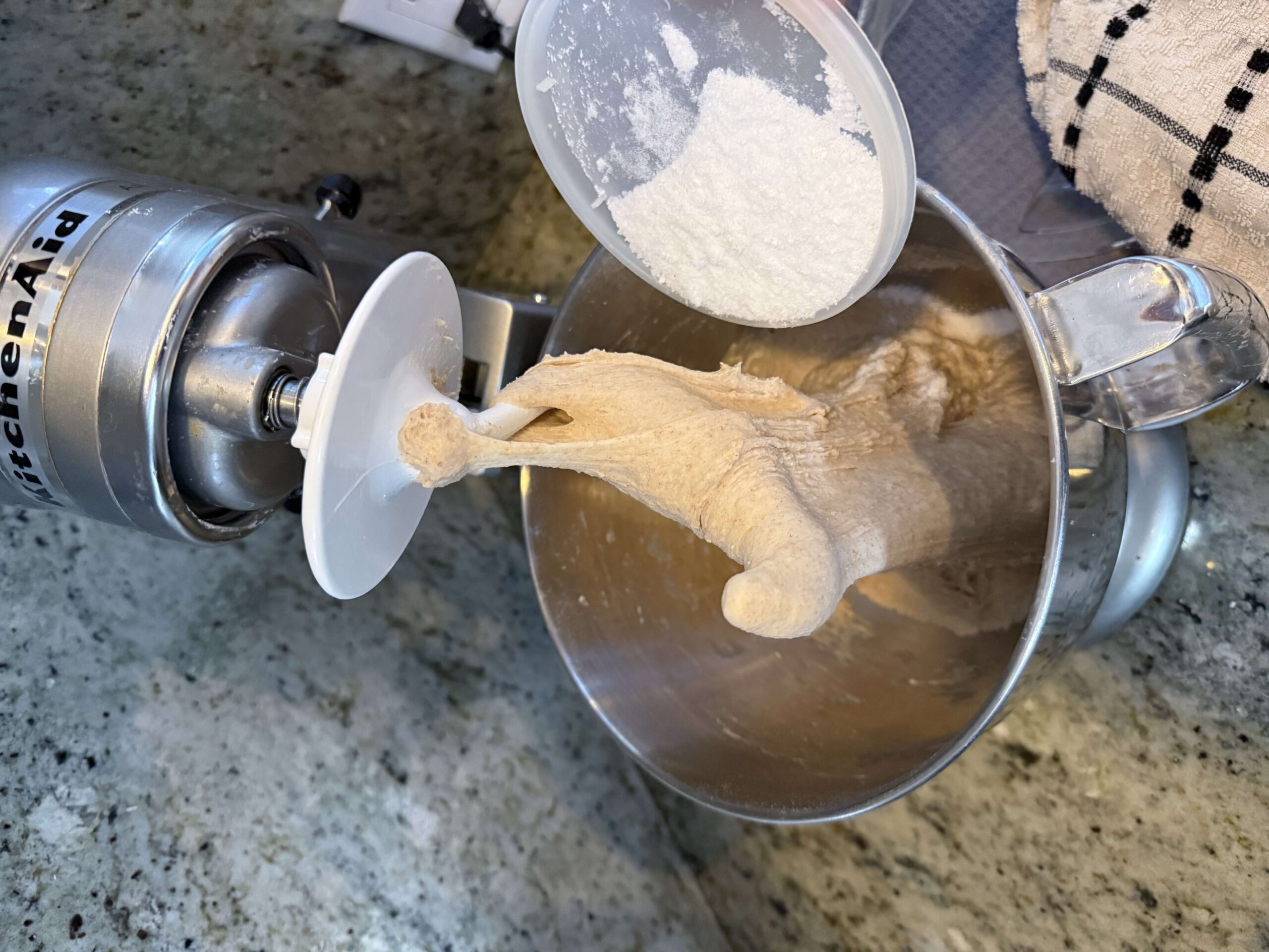 Flour being added to a stand mixer bowl with dough on the hook