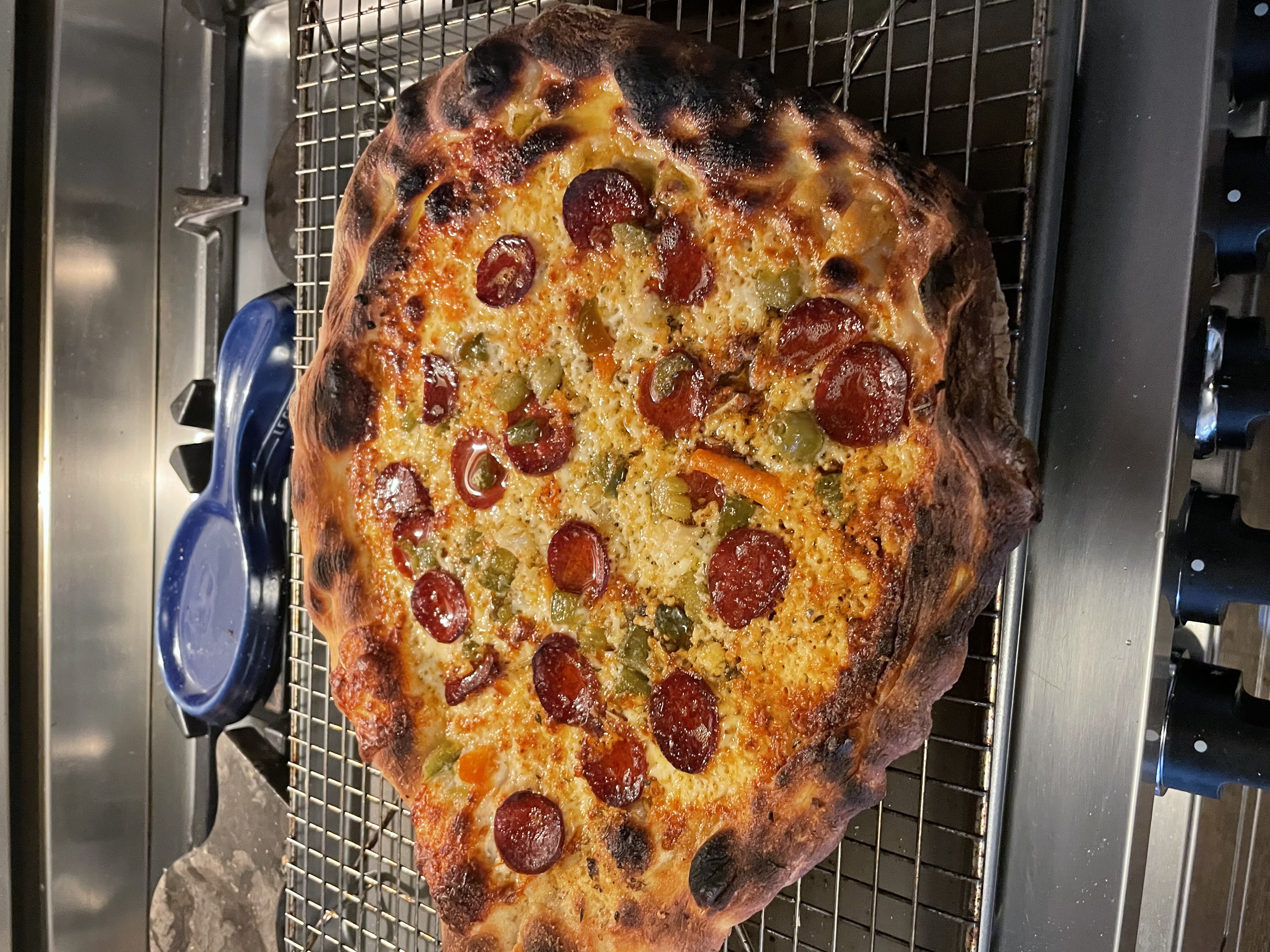 Deeply charred pepperoni and giardiniera pizza cooling on a wire rack after a home oven bake